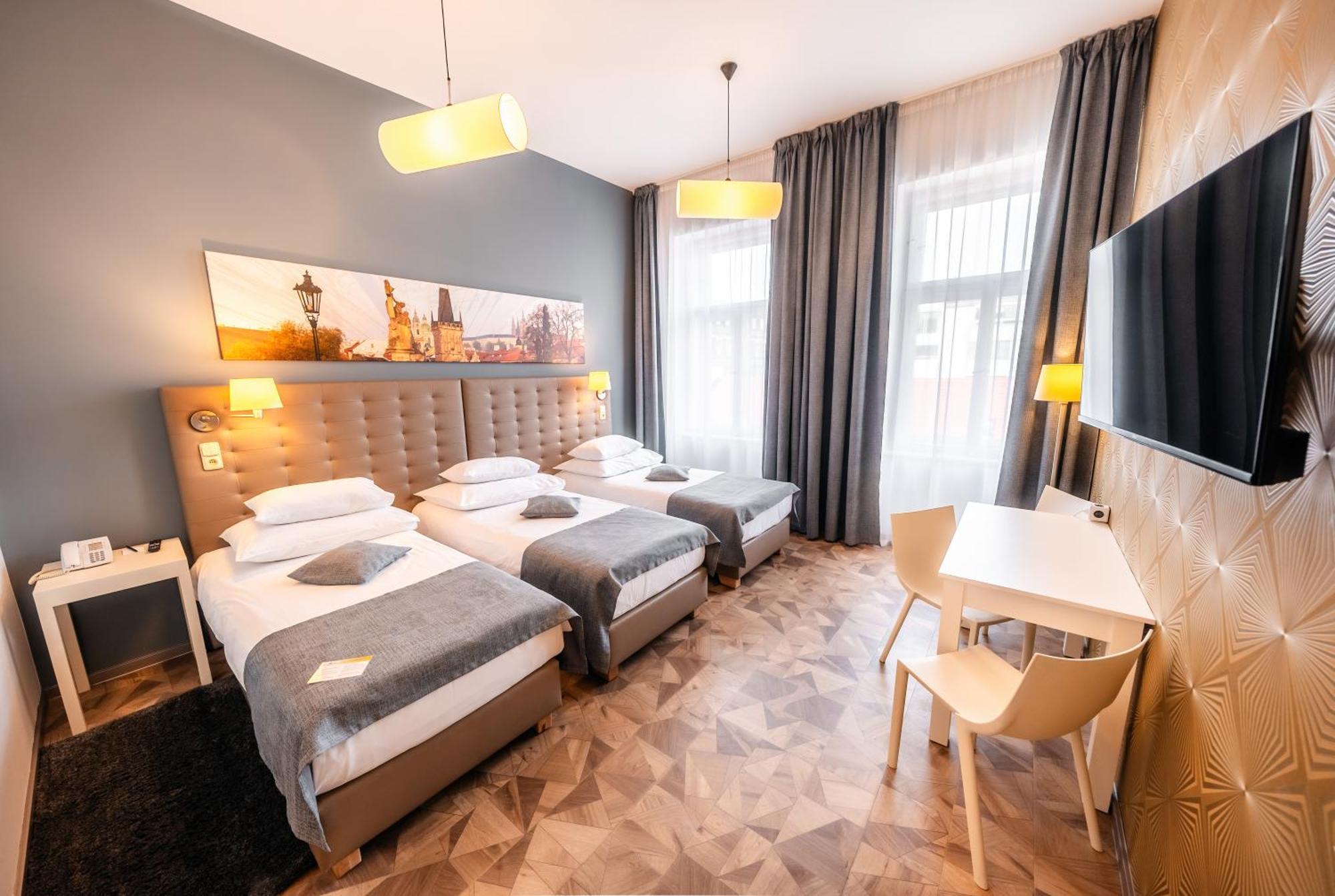 Grandium Hotel Prague Exterior photo The photo shows a modern hotel room featuring three single beds, each with white bedding and gray throws. The walls are painted gray, and artwork depicting a cityscape is displayed above the headboard. There are two stylish pendant lights hanging fro