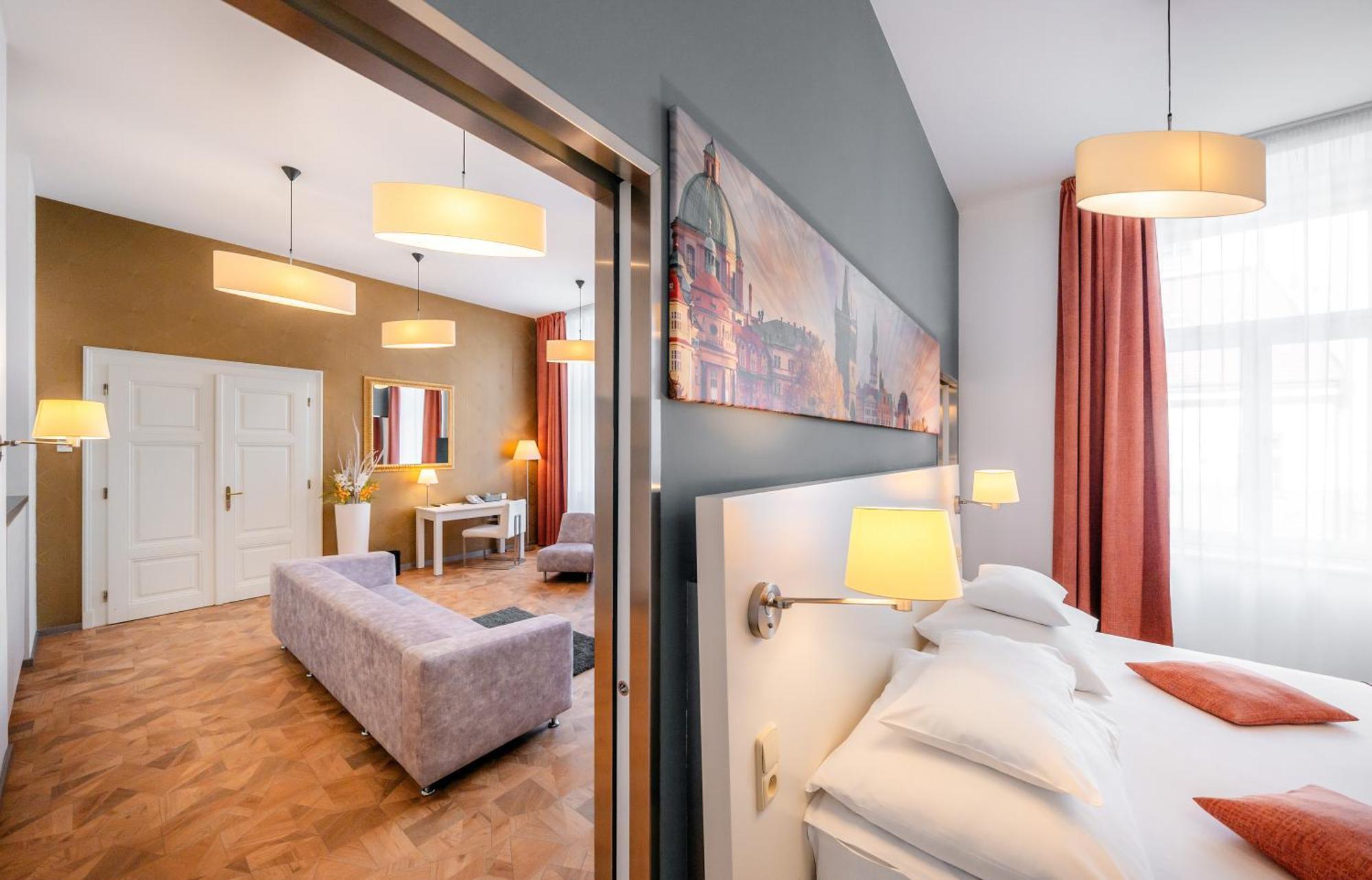 Grandium Hotel Prague Exterior photo The photo shows a stylish hotel room with modern decor. In the foreground, there is a bed with white pillows, and wall-mounted lamps provide soft lighting. Behind the bed, there's an open doorway leading to a living area featuring a light-colored sof